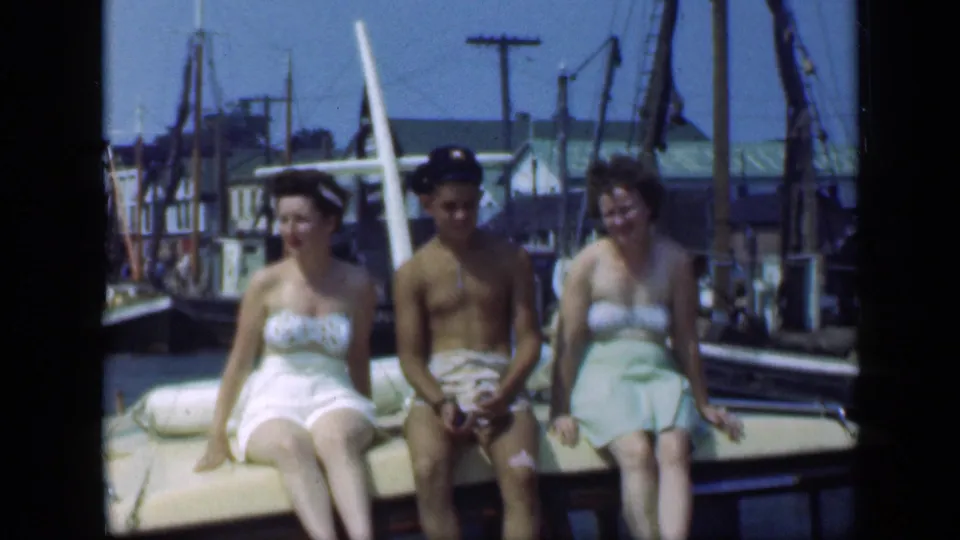 The 1948 Long Island marina scene shows three adults seated on a boat, with swimwear, a sundress, masts, and buildings visible.