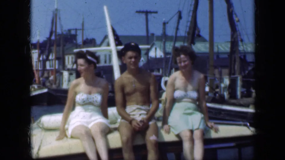 At a Long Island marina in 1948, the three adults continue sitting on the boat as the home-movie camera records the waterfront setting.