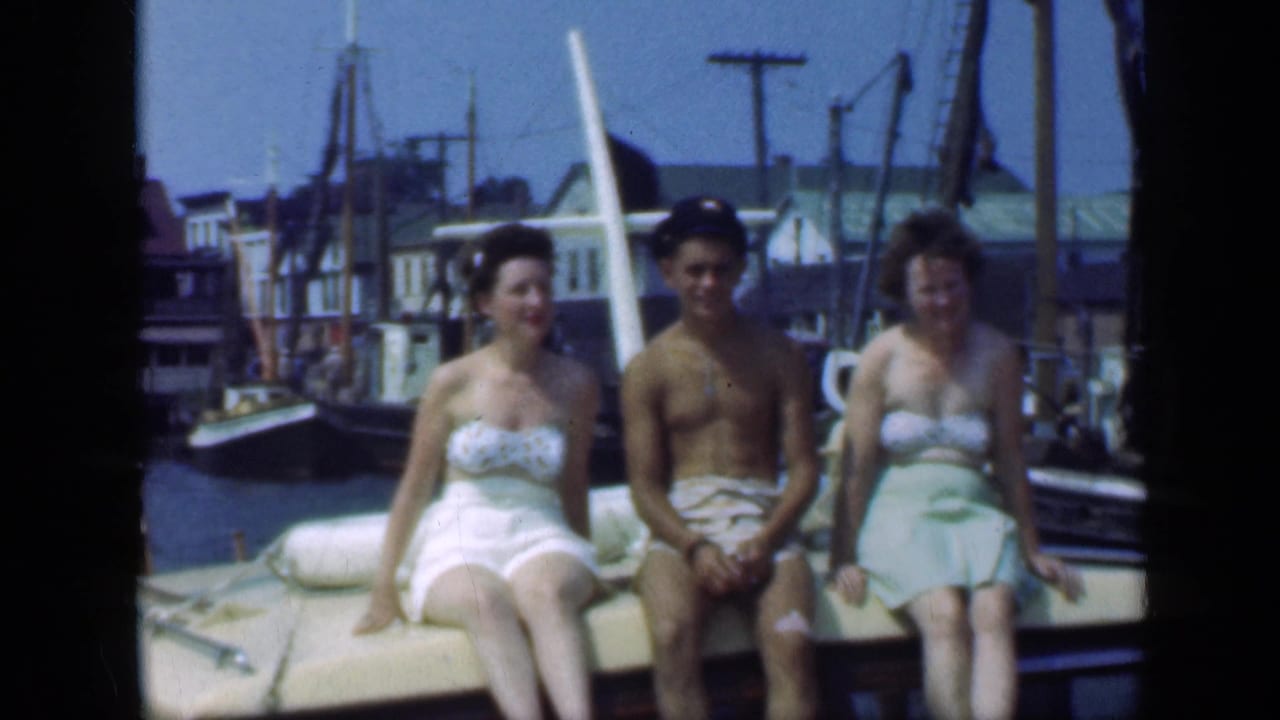Three adults in swimwear sit on a boat with masts and buildings behind them.