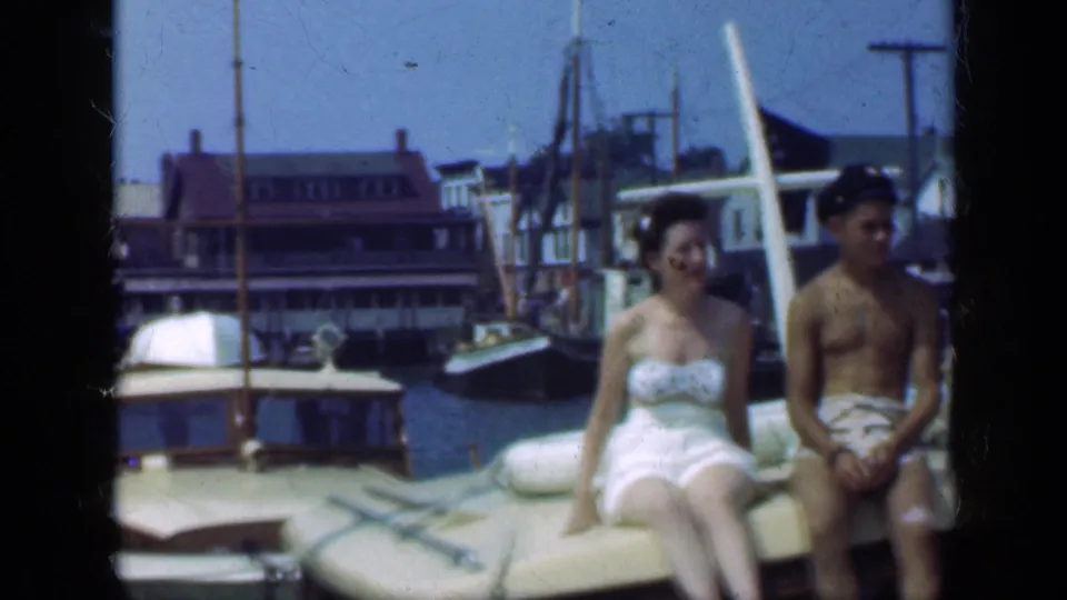 A woman and man sit on a cushioned boat bench in Long Island harbor, with another boat, masts, and waterfront buildings visible behind them.