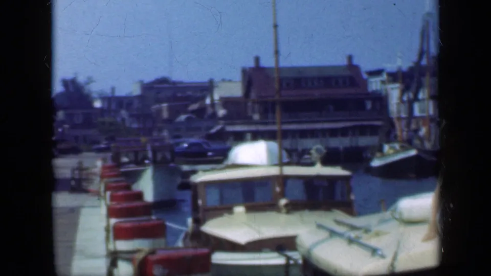 The Long Island harbor view shows moored boats, red dock seating, masts, and waterfront buildings, with part of a seated person at the right edge.