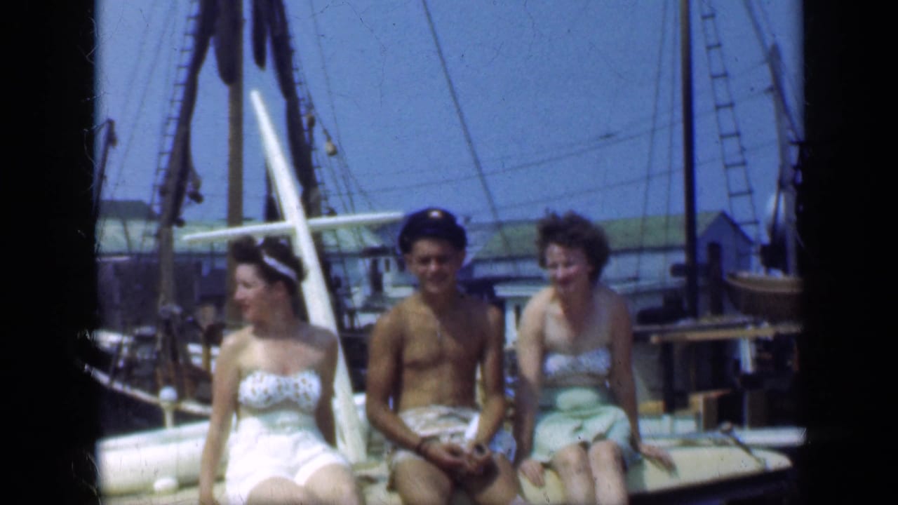 Three adults sit on a sailboat deck in swimwear with marina rigging behind them.