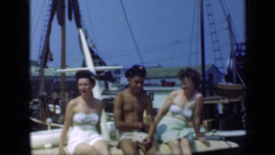 Two women and a man sit close together on a boat deck in Long Island, with rigging, dock buildings, and other boats in the background.