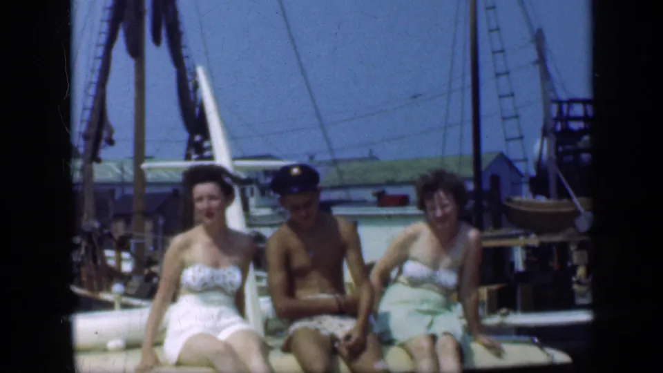 Three adults in summer swimwear sit in a row on the back of a boat at a Long Island marina, framed by rigging and dockside structures.