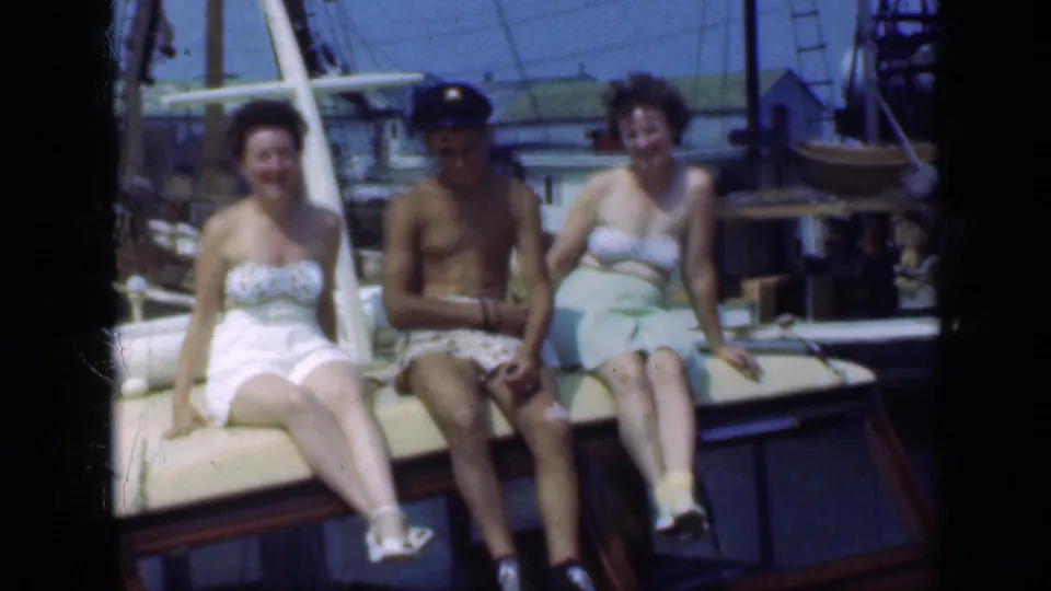 Three adults in swimwear appear seated on a sailboat in Long Island, New York, with masts and nearby boats visible in the background.