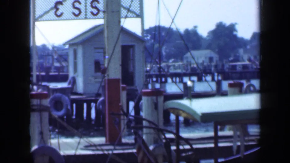 1948 Long Island 8mm frame looking across a marina, with sailboat rigging, a dock, small shore building, moored boat, water, and trees.