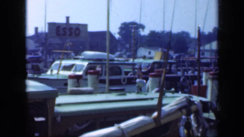 1948 Long Island bay view showing green docked boats, pier posts, mast lines, shoreline buildings, and a distant Esso sign.