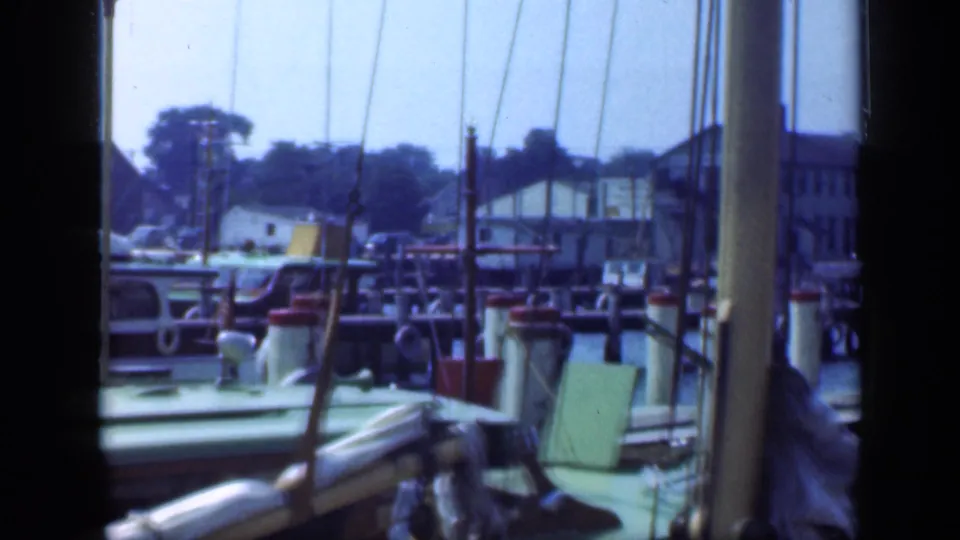 Long Island waterfront frame with a green-roofed boat, dock posts, rigging lines, and houses along the far shore.