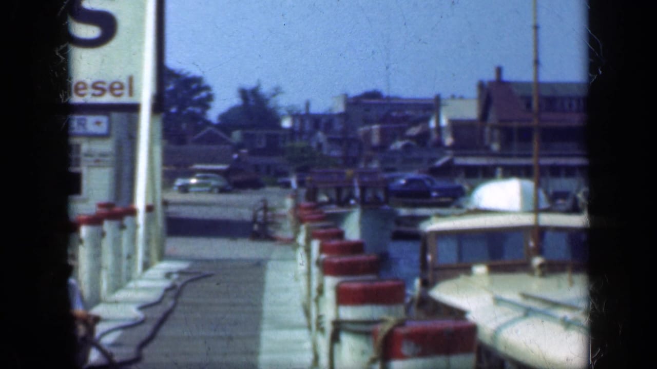 Dock beside moored boats with a marina sign and older cars in the background.
