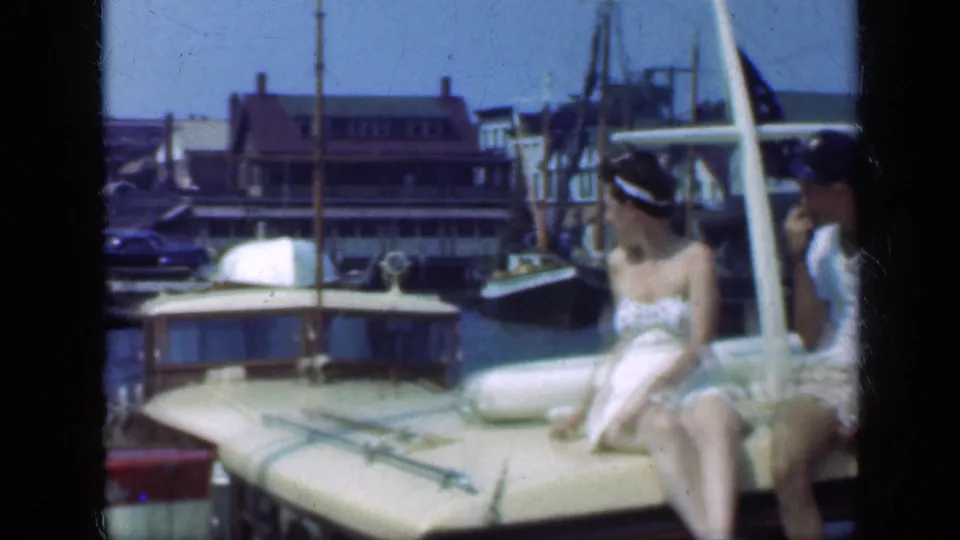 Two women in light summer clothing sit on a moored boat at a 1948 Long Island harbor, with docks and buildings behind them.