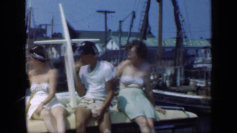 Three adults sit casually on a moored boat in a 1948 Long Island harbor, with cranes, docks, and boat equipment in the background.