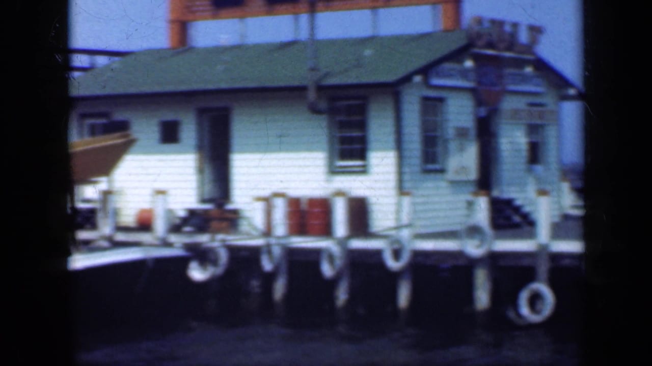 White pier building with green roof beside dock pilings and hanging life rings.