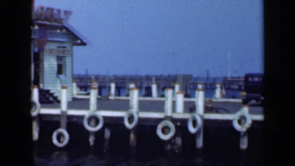 1948 Long Island waterfront view with dock pilings, life rings, calm blue water, distant shoreline, and a dark car near the pier.