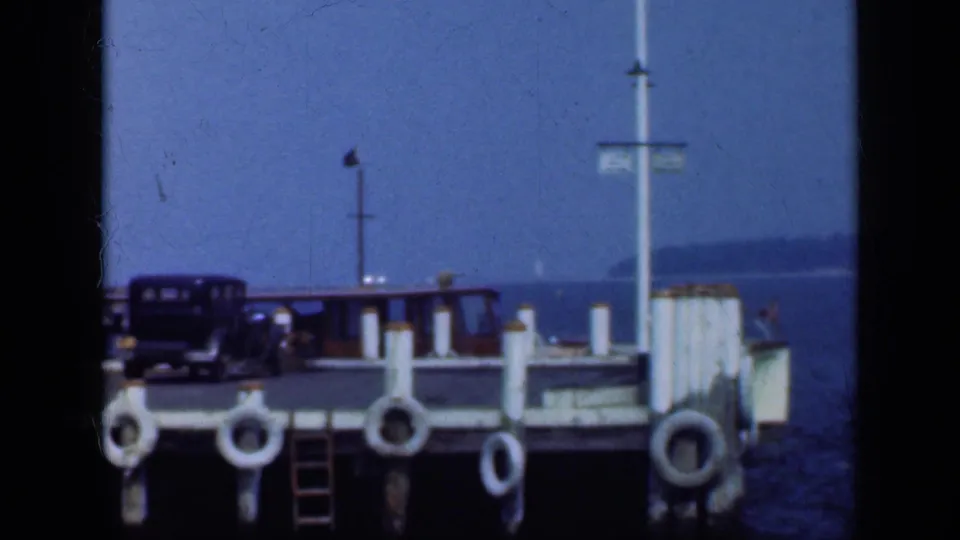 1948 Long Island pier scene with dark automobiles, hanging life rings, a ladder, a vertical signpost, dock rail, water, and shoreline haze.