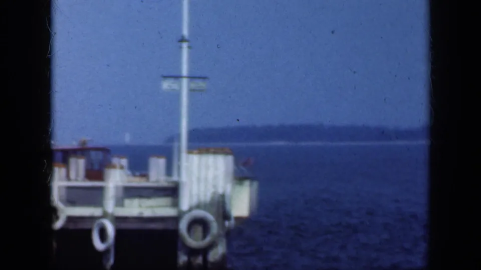 1948 Long Island waterfront frame showing the pier end, hanging life rings, a signpost, calm water, pale sky, and distant shoreline.