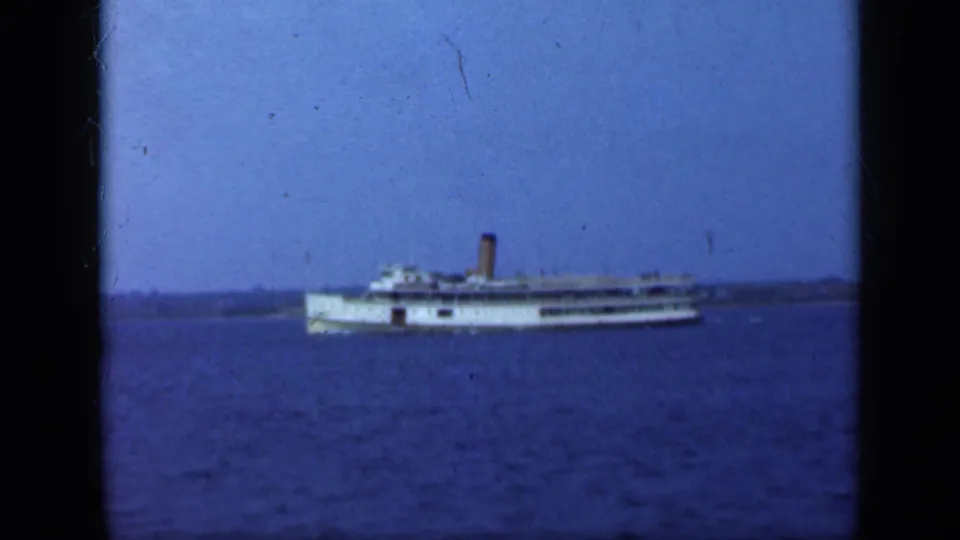 The white steamship remains centered offshore, with its smokestack visible against the blue sky.