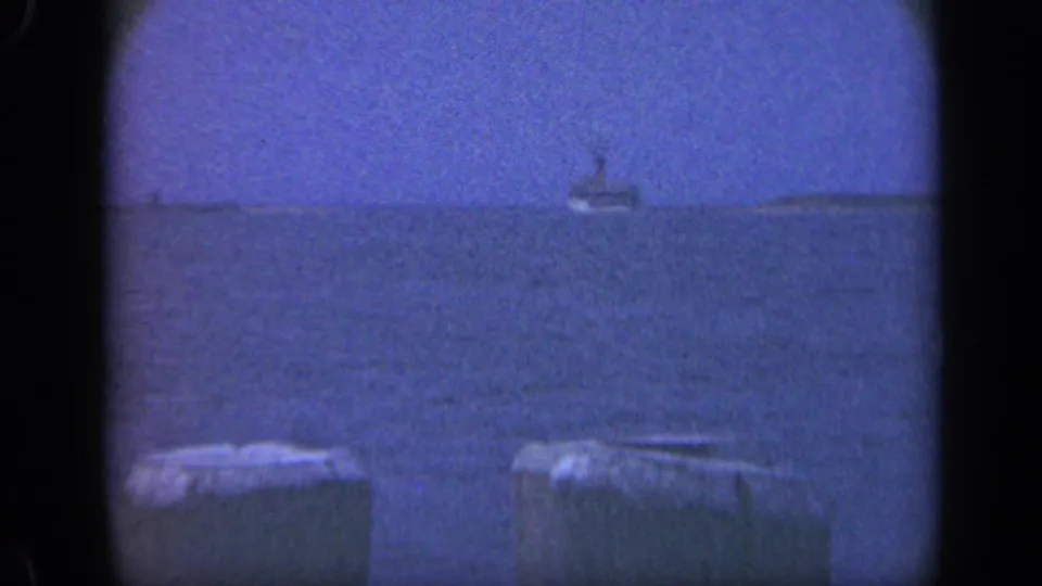 Blue 8mm frame of a large offshore vessel viewed across dark water from a Long Island pier or waterfront edge.