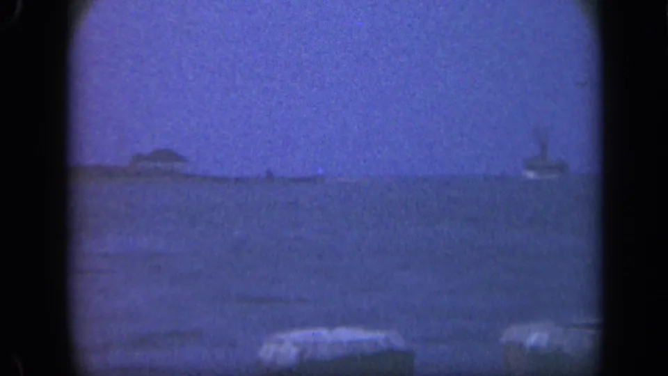 1948 Long Island coastal frame with blue water, distant vessel or shoreline forms, and a dark foreground pier edge.