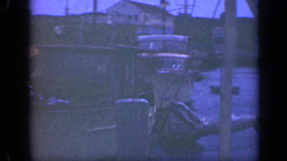 Blue-tinted 8mm frame of small boats tied near dock pilings with a modest shoreline building in the background.