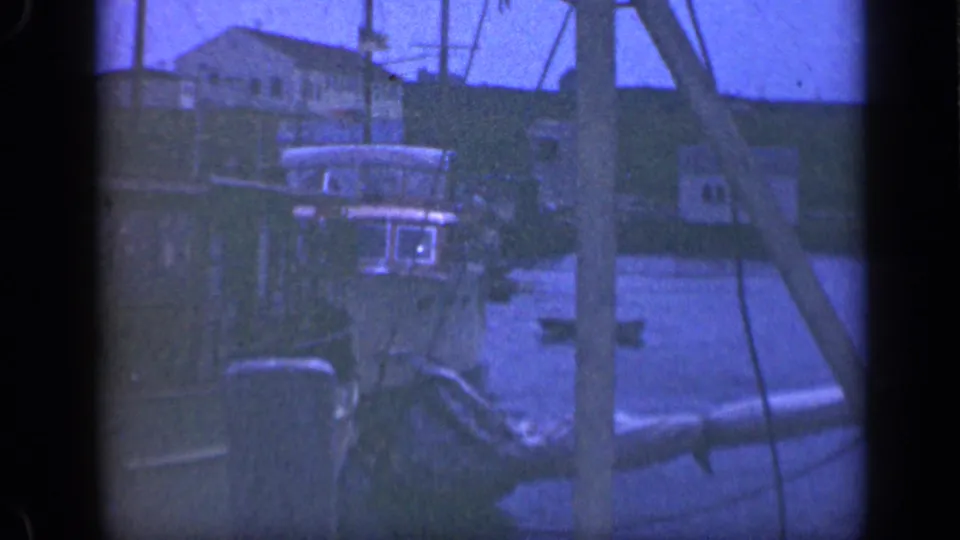 Long Island harbor frame from 1948 showing a small vessel cabin, dock posts, rigging, and houses along the waterfront.