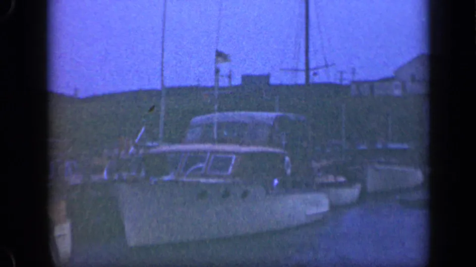 A 1948 Long Island harbor view with a cabin boat in the foreground, sailboats nearby, masts, flags, and low buildings beyond the water.