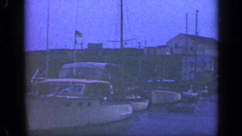 Blue-tinted 8mm frame of docked boats on Long Island, with a cabin cruiser, sailboat masts, flagpoles, and waterfront buildings visible.