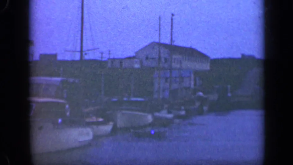 A 1948 Long Island waterfront frame shows small docked boats, masts, a flagpole, bay water, and a shoreline building in soft 8mm focus.