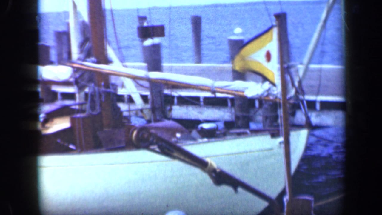 Docked sailboat with rigging, covered boom, pilings, blue water, and a small yellow pennant.