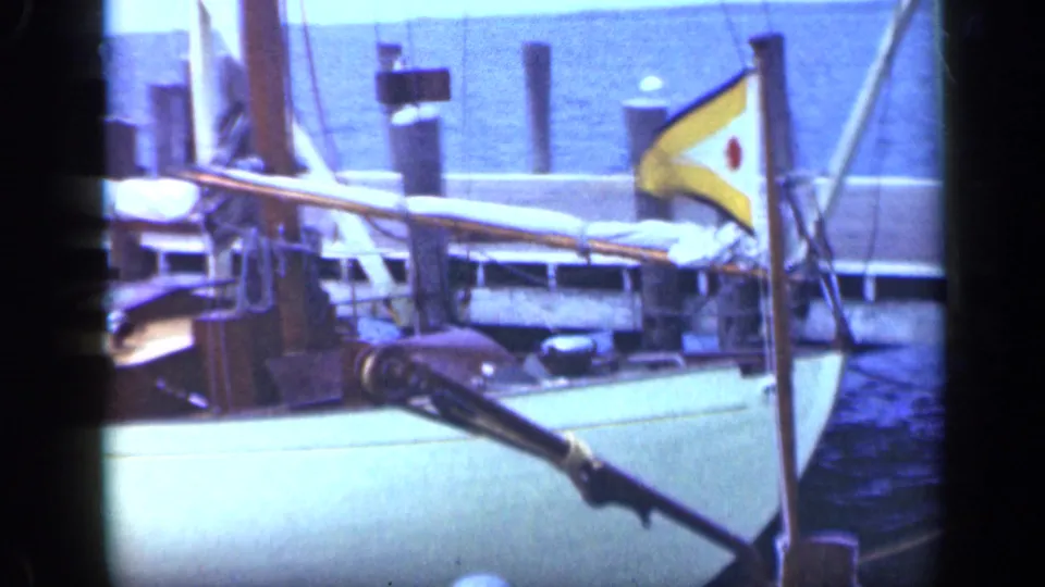 1948 Long Island harbor view of a docked sailboat with rigging, covered boom, dock pilings, blue water, and a yellow pennant.