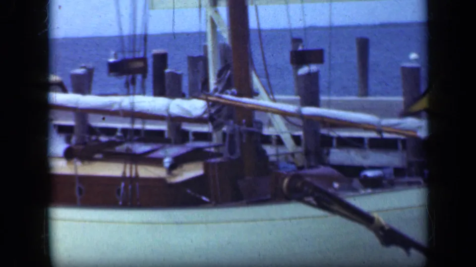 1948 Long Island sailboat detail with wooden deck, covered boom, rigging lines, harbor pilings, and water beyond.