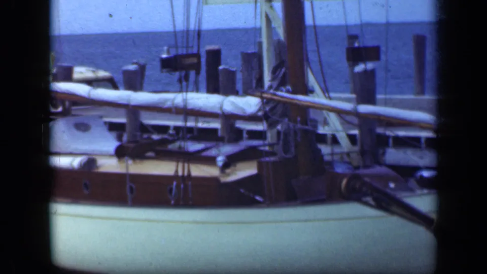 1948 Long Island dockside frame showing sailboat hull, wooden deck, covered boom, rigging, pilings, and harbor water.