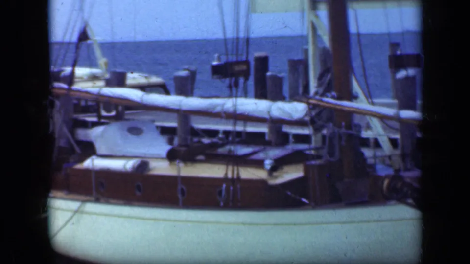 1948 Long Island harbor shot of a docked sailboat with pale hull, cabin ports, covered boom, rigging, and blue water beyond.