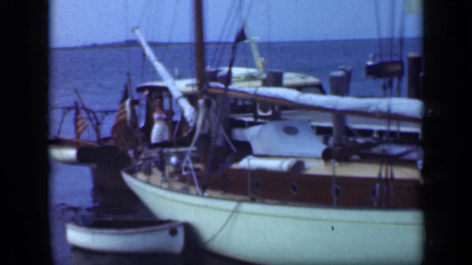 1948 Long Island home-movie frame showing adults on a white sailboat, with a dinghy in front and mast rigging against open water.