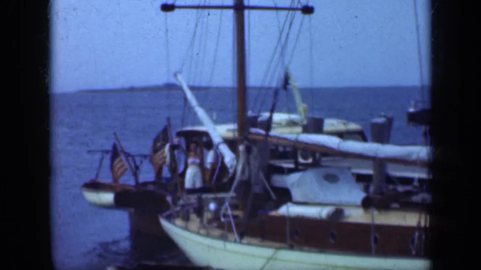 1948 Long Island 8mm frame showing wooden sailboats at a dock, American flags, water, and people aboard the vessels.