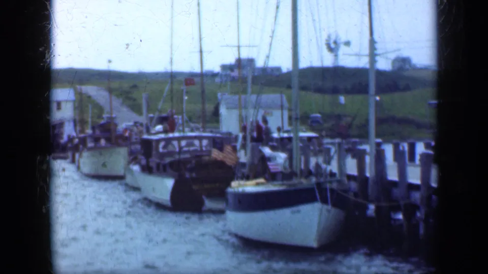 1948 Long Island harbor view with moored boats, tall masts, a wooden pier, grassy bank, and small buildings in the distance.