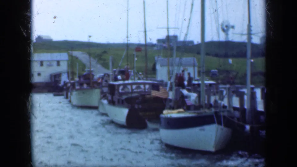 1948 Long Island 8mm frame of sailboats tied at a pier, with people near the boats and a road or path rising through grass behind them.