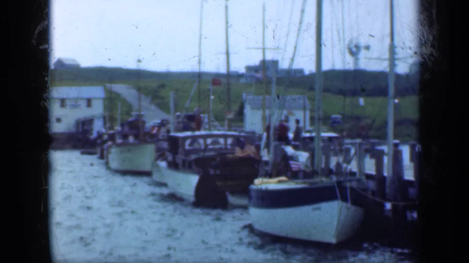 1948 Long Island harbor frame showing docked sailboats, rigging, pier posts, water, grassy shore, and small waterfront buildings.