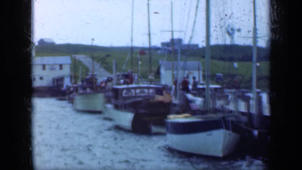 1948 Long Island harbor scene with white boats moored at a wooden pier, sail masts, water, grassy banks, and simple buildings in the background.