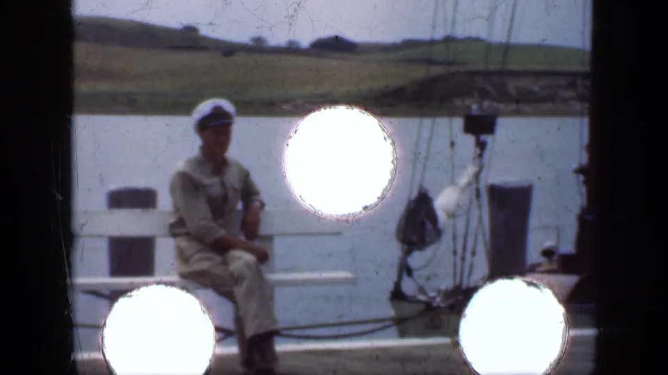 A man in a light outfit and cap sits near a docked boat on the Long Island waterfront, with water, rigging, and circular contact-sheet marks visible.