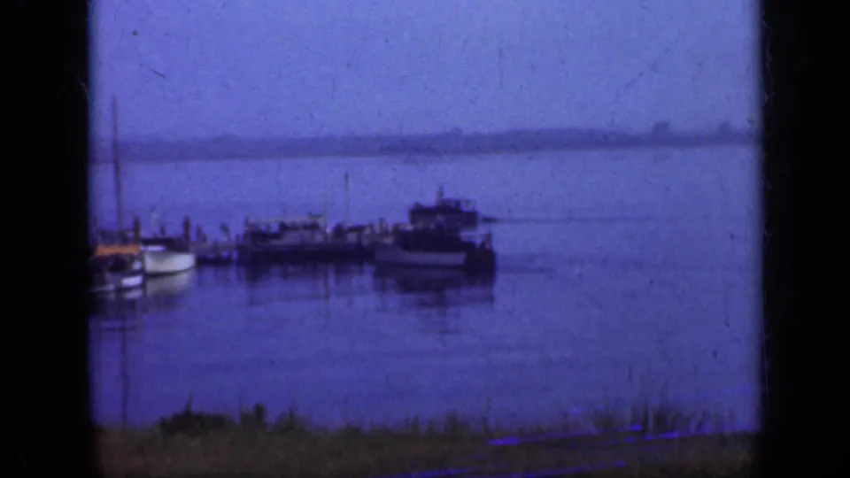 Blue-toned 1948 Long Island harbor view with distant boats tied along a wooden pier, still water, low horizon, and grass in the foreground.
