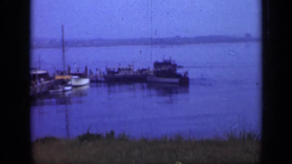 Wide blue-toned Long Island harbor frame showing moored boats, a pier, calm water, foreground grass, and a distant low shoreline.