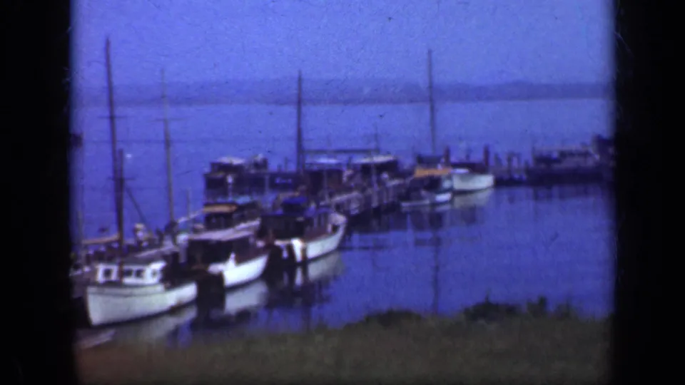 A closer 1948 home-movie view shows moored boats along a wooden dock with blue water and shoreline behind them.