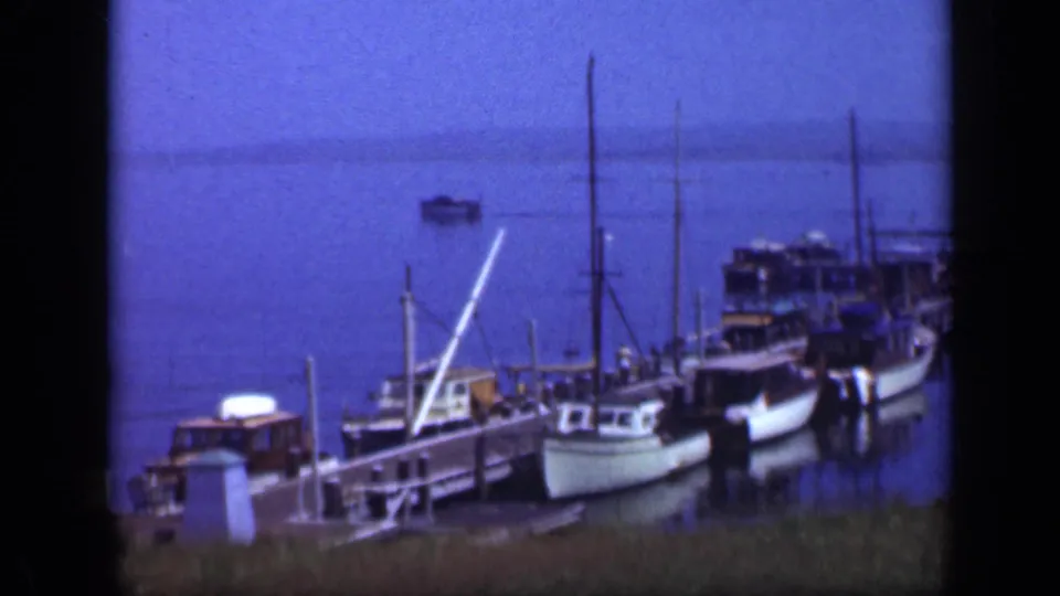 1948 Long Island waterfront frame showing boats docked along a wooden pier, open harbor water, and a distant vessel.