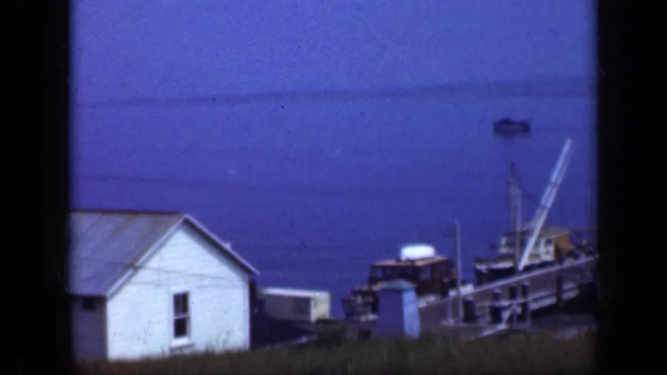1948 Long Island waterfront frame with a small white building, docked boats, pier railings, and calm blue harbor water.