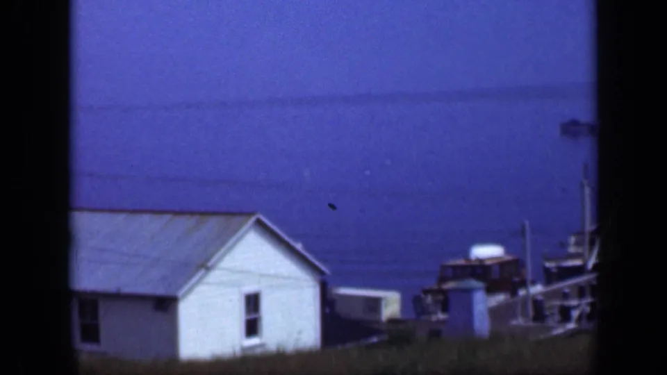 1948 Long Island home-movie frame showing a small white waterside house, blue water, utility lines, grass, and a small boat nearby.
