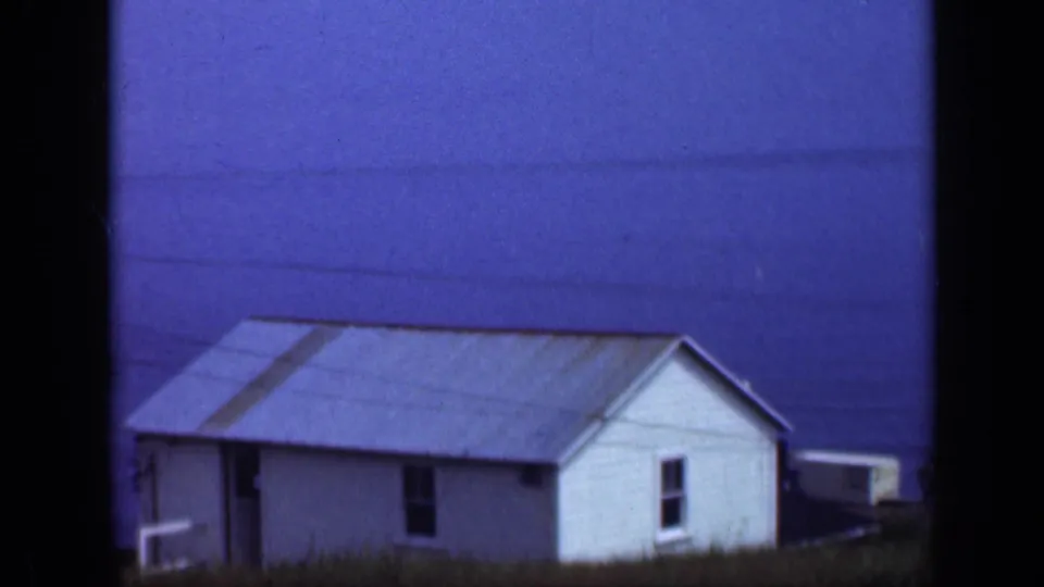 1948 Long Island frame of a simple white coastal house with a weathered roof, dark window openings, and blue water behind it.