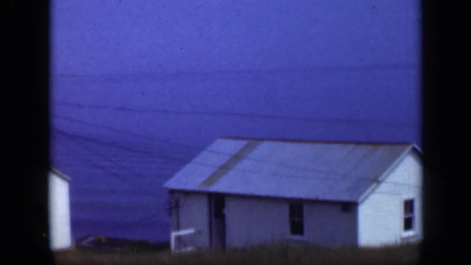 1948 Long Island footage frame showing a low white seaside house, sloped metal roof, blue water, and grassy foreground.