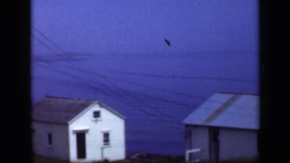 1948 Long Island home-movie frame with two small white waterside buildings, blue water, utility lines, and dark 8mm frame edges.