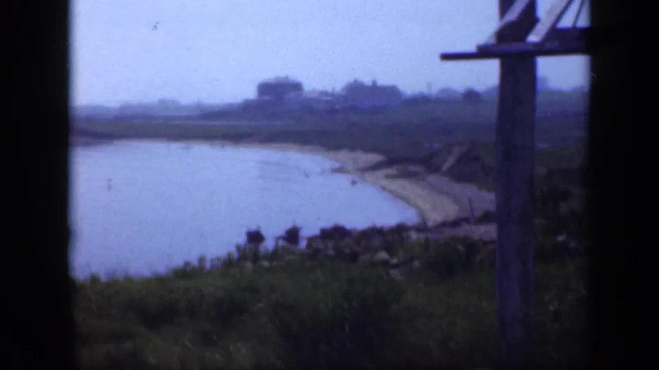 1948 Long Island home-movie frame looking across a sandy cove, grassy foreground, shoreline curve, and soft distant buildings.