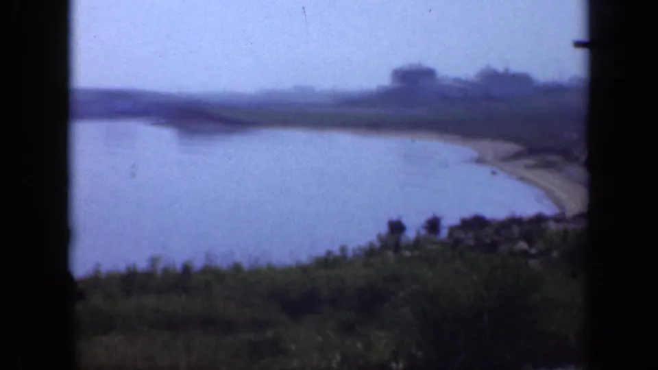 1948 Long Island coastal frame showing calm cove water, a grassy bank, sandy shoreline, and distant buildings softened by 8mm focus.